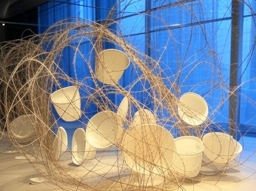 Instalação artística imersiva sobre fragilidade e poder em galeria brasileira, com elementos simbólicos como arame farpado e porcelanas quebradas.